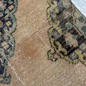 May include: A woven rug featuring a tan background with intricate, symmetrical patterns in shades of black, dark green, and muted yellow. The textile displays a dense, knotted pile construction with visible fiber textures throughout the surface.