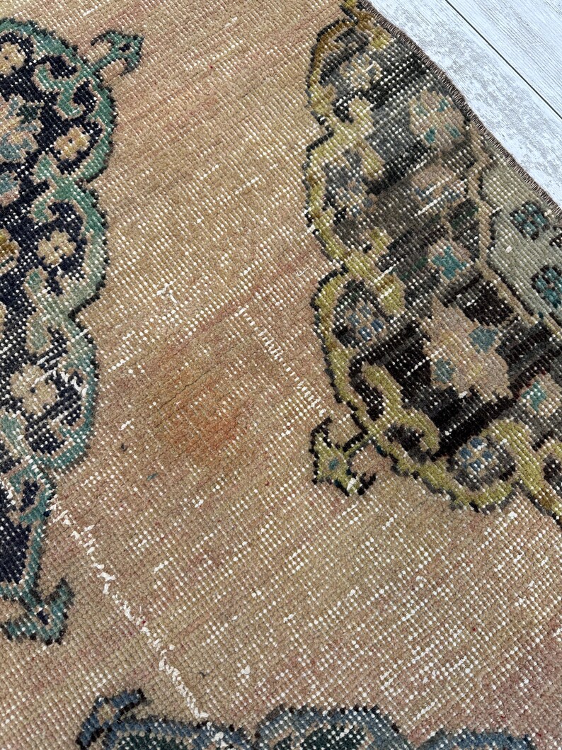 May include: A woven rug featuring a tan background with intricate, symmetrical patterns in shades of black, dark green, and muted yellow. The textile displays a dense, knotted pile construction with visible fiber textures throughout the surface.