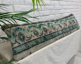 May include: A long, rectangular pillow with a vintage rug design. The pillow features a teal, brown, and cream pattern. The pillow is resting on a white surface, with a white brick wall in the background.