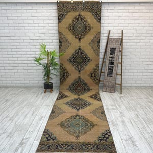 May include: A long, narrow runner rug featuring a tan background with repeating dark blue and teal diamond-shaped medallions. The rug has a dark patterned border at both ends. A wooden ladder displays a sign reading TURKISH RUG & MORE.