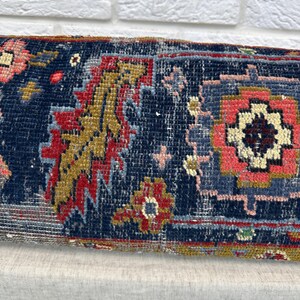 May include: Close-up of a rectangular, patterned textile. The rug features a dark blue background with intricate designs in red, gold, pink, and light blue. The textile has a vintage, worn appearance, suggesting it is an antique or a piece with a distressed aesthetic.