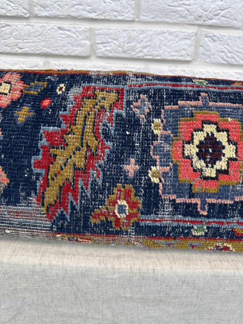May include: Close-up of a rectangular, patterned textile. The rug features a dark blue background with intricate designs in red, gold, pink, and light blue. The textile has a vintage, worn appearance, suggesting it is an antique or a piece with a distressed aesthetic.