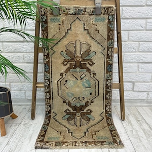 May include: A rectangular, beige rug with a central geometric design in brown and teal. The rug is displayed on a wooden ladder against a white brick wall. The rug has a fringed edge and a detailed border.