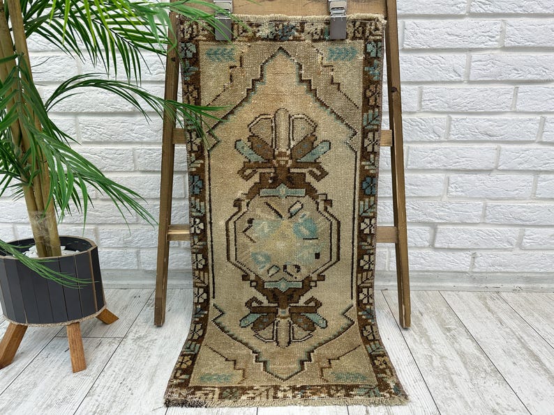 May include: A rectangular, beige rug with a central geometric design in brown and teal. The rug is displayed on a wooden ladder against a white brick wall. The rug has a fringed edge and a detailed border.