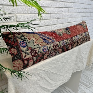 May include: A long, rectangular pillow with a vintage rug design. The pillow features a red central field with blue, beige, and pink geometric patterns. The edges are dark with intricate floral motifs. The pillow is placed on a white surface.