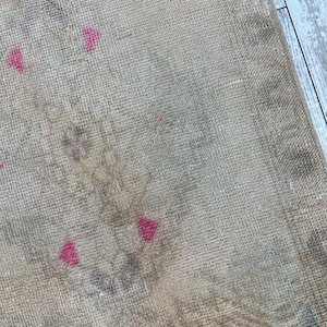 K&ouml;nnte beinhalten: Ein gewebter Teppich mit einer neutralen beigen Basis und einer verblassten, abgenutzten Textur. Die Oberfl&auml;che zeigt dezente, ged&auml;mpfte graue Blumenmuster und kleine, markante geometrische Akzente in einem leuchtenden Pink.
