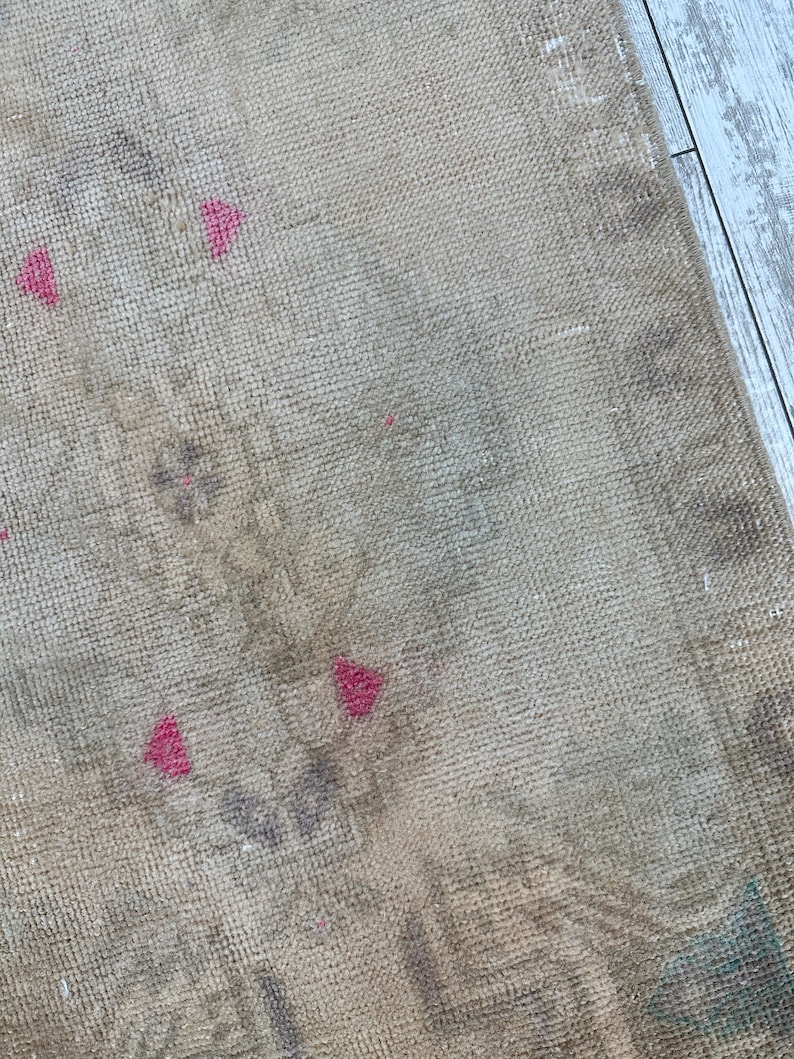K&ouml;nnte beinhalten: Ein gewebter Teppich mit einer neutralen beigen Basis und einer verblassten, abgenutzten Textur. Die Oberfl&auml;che zeigt dezente, ged&auml;mpfte graue Blumenmuster und kleine, markante geometrische Akzente in einem leuchtenden Pink.