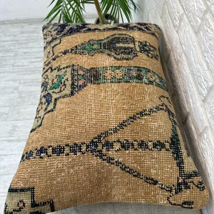 May include: Beige rectangular cushion with a woven, patterned design. It features a central band of green, black, and blue geometric shapes, with additional blue and pink accents. The cushion is placed against a white brick wall.