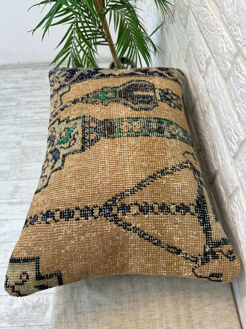 May include: Beige rectangular cushion with a woven, patterned design. It features a central band of green, black, and blue geometric shapes, with additional blue and pink accents. The cushion is placed against a white brick wall.