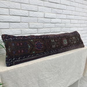 May include: A long, rectangular lumbar pillow featuring a dark, multi-colored woven textile pattern. The design includes intricate geometric and floral motifs in shades of deep brown, navy blue, muted red, and soft green across the entire surface.