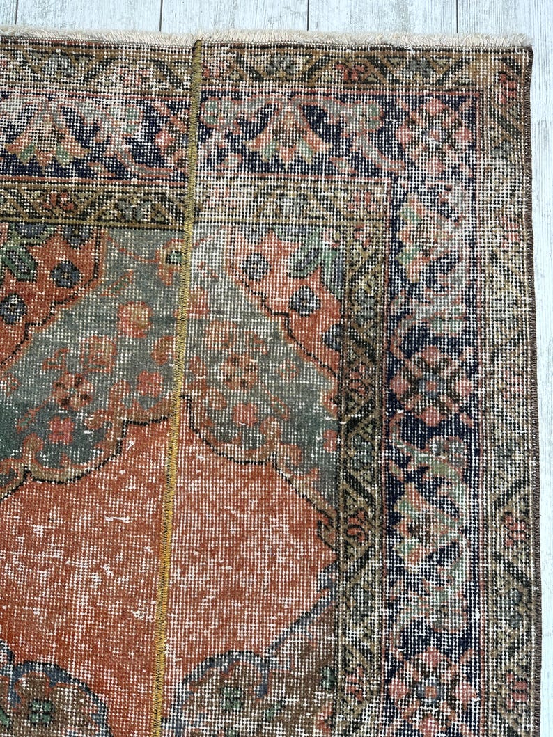 May include: A rectangular area rug featuring a central field of muted orange with intricate floral and geometric patterns in shades of sage green, navy blue, and cream. The rug has a wide, detailed border and a short, light-colored fringe edge.
