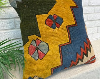 May include: A square throw pillow featuring a woven geometric pattern in dark green, mustard yellow, and blue. The design includes zigzag lines and stylized floral motifs in red, blue, and light gray, with a textured, coarse fabric construction.
