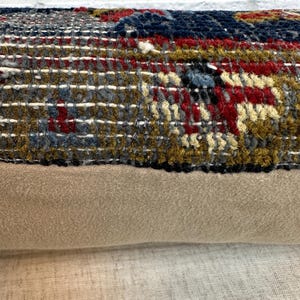 May include: Close-up of a decorative pillow with a woven, textured top in shades of gray, red, blue, and gold. The bottom portion is a solid tan fabric. The pillow is likely for home decor.