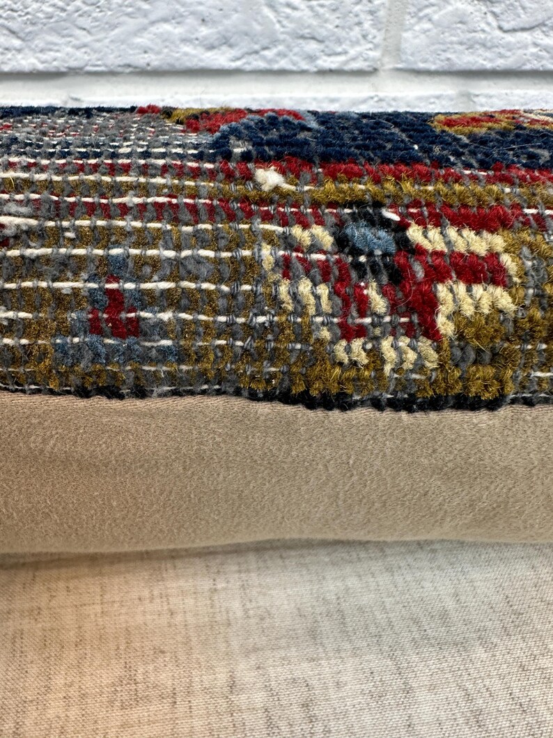 May include: Close-up of a decorative pillow with a woven, textured top in shades of gray, red, blue, and gold. The bottom portion is a solid tan fabric. The pillow is likely for home decor.