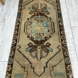 May include: A rectangular, vintage rug with a central geometric design in shades of brown, beige, and teal. The rug features a detailed border and a distressed appearance, suggesting age and use. The rug is placed on a light-coloured wooden floor.