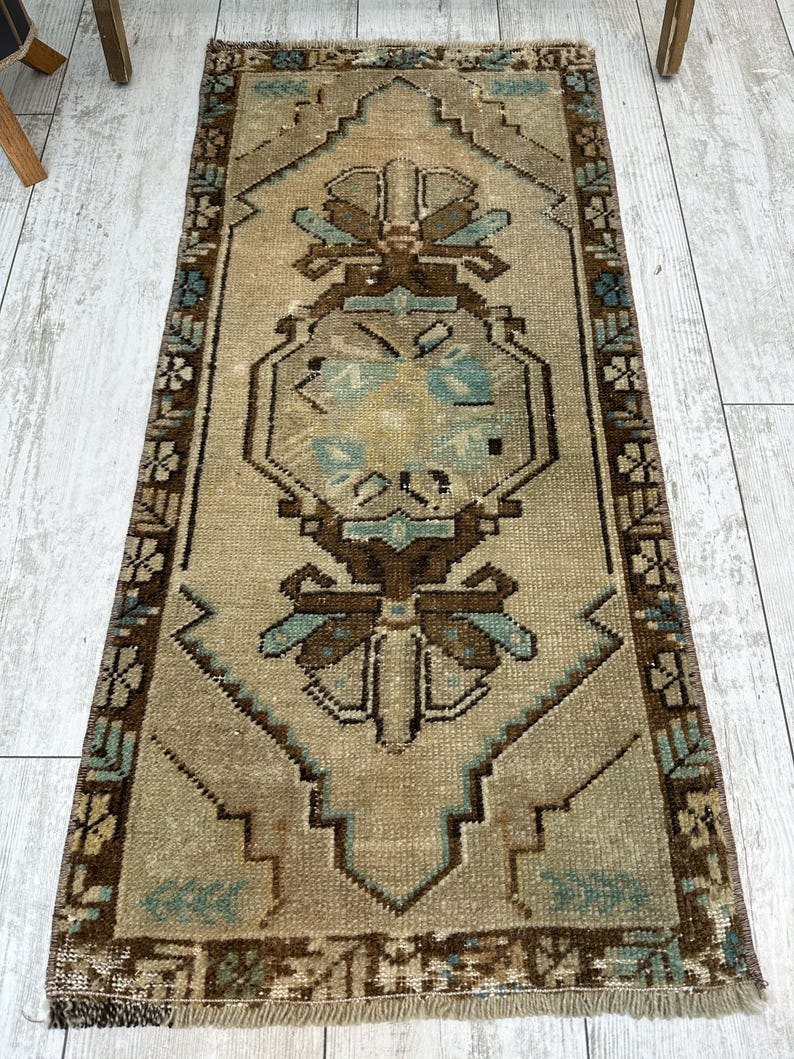 May include: A rectangular, vintage rug with a central geometric design in shades of brown, beige, and teal. The rug features a detailed border and a distressed appearance, suggesting age and use. The rug is placed on a light-coloured wooden floor.