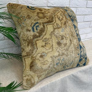 May include: A square throw pillow featuring a woven textile cover with a distressed, vintage-inspired pattern in shades of tan, beige, and brown, accented by a prominent vertical border of deep blue geometric and floral motifs across the right side.