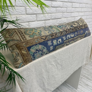 May include: A long rectangular lumbar pillow featuring a woven textile pattern with a distressed appearance. The design incorporates geometric and floral motifs in shades of deep blue, muted brown, beige, and soft teal, creating a textured look.