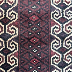 May include: Close-up of a patterned rug with a repeating design. The rug features a central burgundy panel with diamond and geometric shapes, flanked by panels with cream and dark brown scrollwork. The rug has a textured appearance.