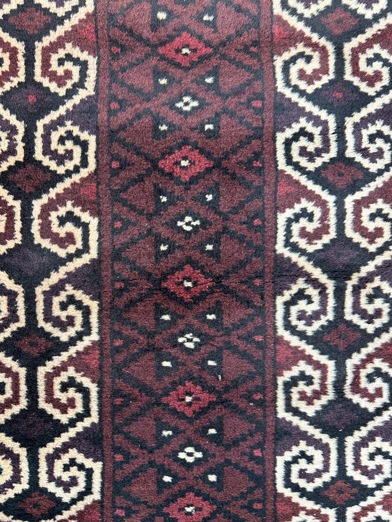 May include: Close-up of a patterned rug with a repeating design. The rug features a central burgundy panel with diamond and geometric shapes, flanked by panels with cream and dark brown scrollwork. The rug has a textured appearance.