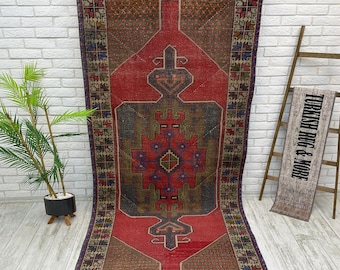 May include: A rectangular, vintage rug with a central red field and intricate geometric patterns in shades of green, brown, and blue. The rug has a border with repeating floral motifs. The rug is hanging against a white brick wall.