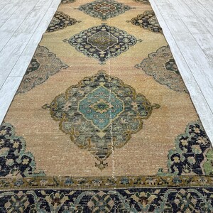 May include: A long, rectangular runner rug featuring a tan base color with a repeating series of large, ornate diamond-shaped medallions in shades of teal, navy blue, olive green, and cream, finished with a dark, patterned border.