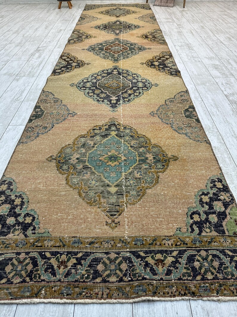 May include: A long, rectangular runner rug featuring a tan base color with a repeating series of large, ornate diamond-shaped medallions in shades of teal, navy blue, olive green, and cream, finished with a dark, patterned border.