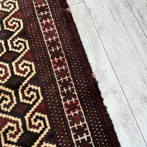 May include: Close-up of a section of a rug with a geometric pattern. The rug features a dark red and cream color scheme with intricate designs. The border has a repeating pattern of red and white shapes.