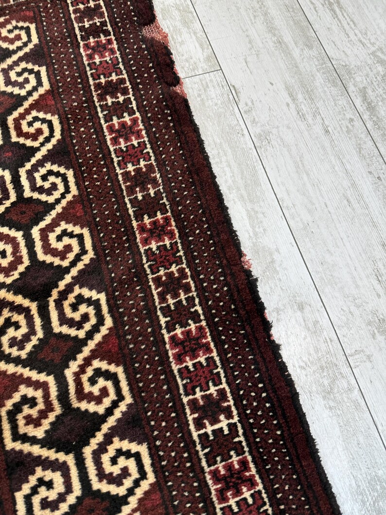 May include: Close-up of a section of a rug with a geometric pattern. The rug features a dark red and cream color scheme with intricate designs. The border has a repeating pattern of red and white shapes.