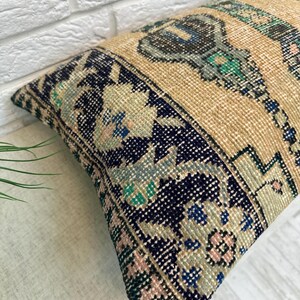 May include: Rectangular beige cushion with a navy blue border featuring floral and geometric patterns in pink, blue, and green. The cushion has a textured, woven appearance, suggesting a vintage or handcrafted style. The central design includes a stylized motif.