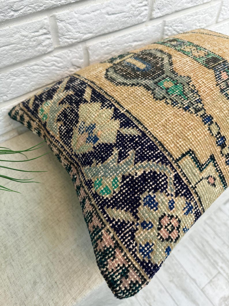 May include: Rectangular beige cushion with a navy blue border featuring floral and geometric patterns in pink, blue, and green. The cushion has a textured, woven appearance, suggesting a vintage or handcrafted style. The central design includes a stylized motif.