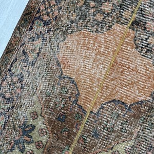 May include: A woven rug featuring a complex pattern with a central orange field surrounded by dark brown, sage green, and black floral and geometric motifs. The textile displays a dense, knotted pile construction with a visible vertical seam.