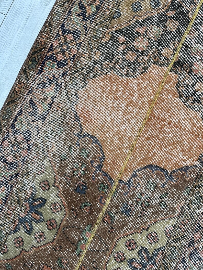 May include: A woven rug featuring a complex pattern with a central orange field surrounded by dark brown, sage green, and black floral and geometric motifs. The textile displays a dense, knotted pile construction with a visible vertical seam.