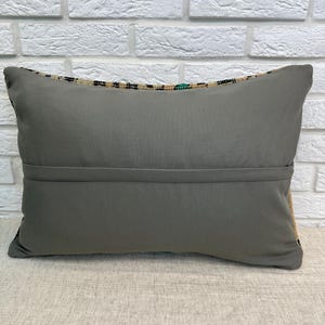 May include: A rectangular grey cushion with a patterned border. The cushion is made of a soft fabric and has a seam running across the middle. The background is a white brick wall.