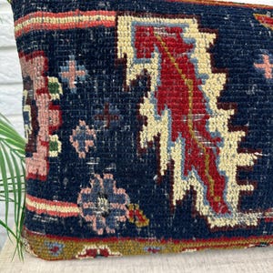 May include: A rectangular pillow with a dark blue background and a detailed, colorful pattern. The design includes red, cream, and light blue geometric shapes, with accents of pink and green. The pillow has a textured, woven appearance.