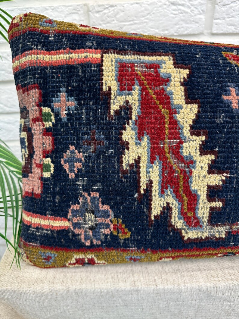 May include: A rectangular pillow with a dark blue background and a detailed, colorful pattern. The design includes red, cream, and light blue geometric shapes, with accents of pink and green. The pillow has a textured, woven appearance.