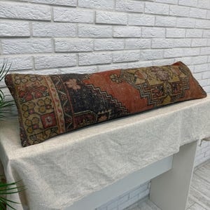 May include: A long, rectangular lumbar pillow featuring a vintage-style woven textile pattern. The design displays a central geometric medallion in shades of burnt orange, deep charcoal, and muted gold, with intricate border details throughout.