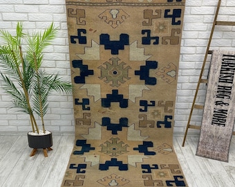 May include: A rectangular runner rug featuring a tan base color with geometric patterns in dark blue and light beige. The design includes repeating symmetrical motifs and angular shapes arranged in a vertical orientation across the entire surface.