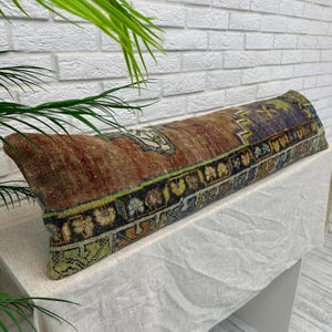 May include: A long, rectangular lumbar pillow featuring a distressed, woven textile cover with a multi-colored geometric pattern in shades of rust brown, deep blue, olive green, and cream, finished with a detailed border of repeating motifs.