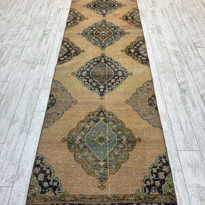 May include: A long, narrow runner rug featuring a tan background with a repeating series of large, ornate diamond-shaped medallions in shades of dark blue, teal, and muted red, finished with a dark blue patterned border at the ends.