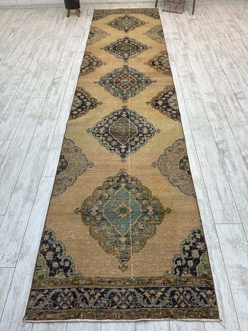 May include: A long, narrow runner rug featuring a tan background with a repeating series of large, ornate diamond-shaped medallions in shades of dark blue, teal, and muted red, finished with a dark blue patterned border at the ends.