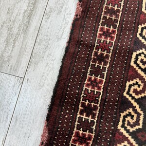 May include: Close-up of a section of a maroon and cream-colored rug. The rug features a geometric pattern with a central border of repeating shapes and a dotted background. The rug's texture is visible, with a slightly frayed edge.