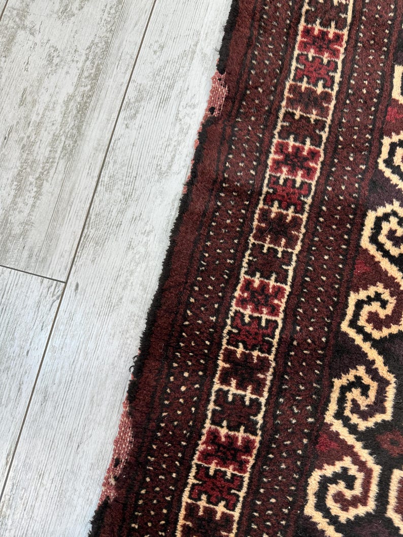 May include: Close-up of a section of a maroon and cream-colored rug. The rug features a geometric pattern with a central border of repeating shapes and a dotted background. The rug's texture is visible, with a slightly frayed edge.