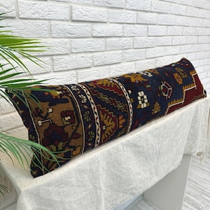 May include: A long, rectangular pillow with a patterned design. The pillow features a dark blue background with intricate patterns in shades of brown, red, and gold. The pillow is placed on a white surface against a white brick wall.