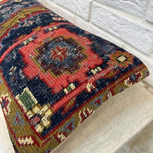 May include: A rectangular pillow with a detailed, colorful rug design. The pillow features a navy blue background with red, green, and gold geometric patterns. The pillow is resting on a neutral-colored surface.