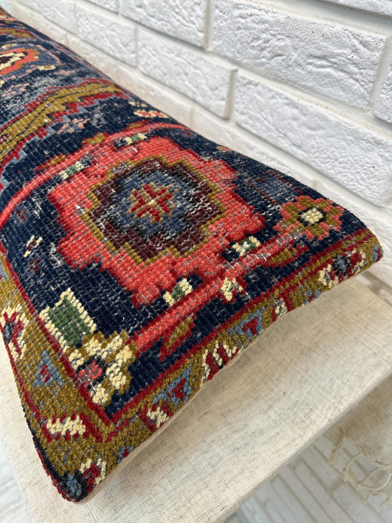 May include: A rectangular pillow with a detailed, colorful rug design. The pillow features a navy blue background with red, green, and gold geometric patterns. The pillow is resting on a neutral-colored surface.