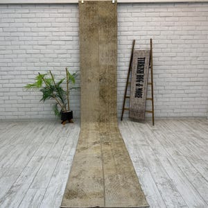 May include: A long, rectangular, beige and brown rug with a distressed, vintage appearance. The rug is displayed on a light wood floor in front of a white brick wall. A wooden ladder with a sign that reads "TURKISH RUG & MORE" is to the right.