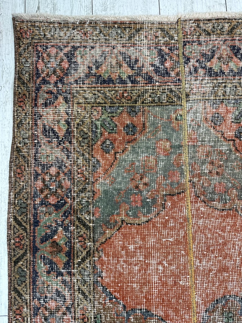 May include: A rectangular area rug featuring a complex pattern with a central field of muted orange, surrounded by borders in shades of navy blue, sage green, and dusty pink. The design includes intricate floral and geometric motifs throughout.