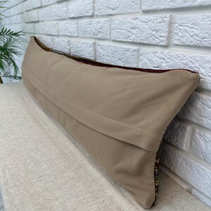May include: A long, rectangular beige pillow with a textured fabric cover. The pillow is placed on a light-colored surface against a white brick wall. The edges of the pillow show a colorful, patterned design.