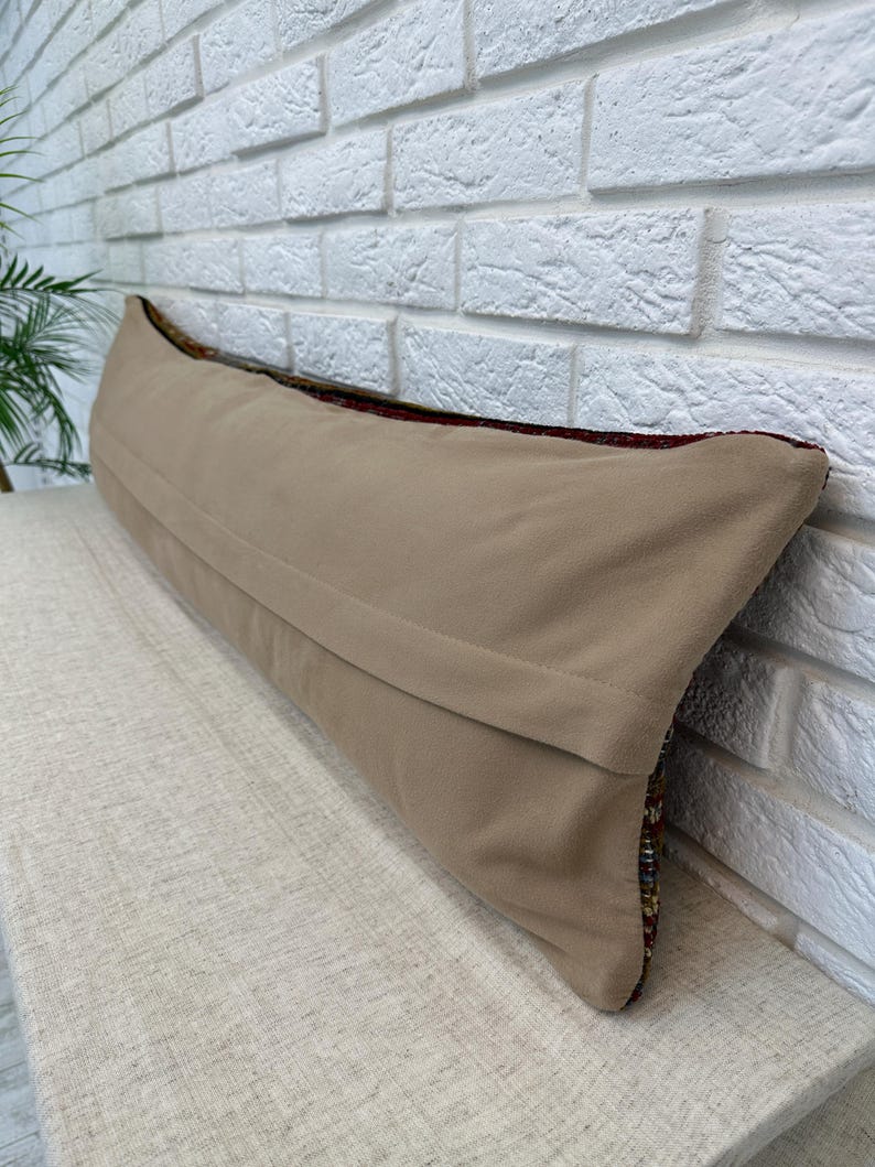 May include: A long, rectangular beige pillow with a textured fabric cover. The pillow is placed on a light-colored surface against a white brick wall. The edges of the pillow show a colorful, patterned design.